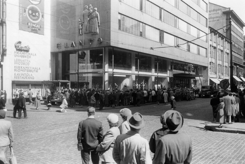 JALANKULKIJOITA ALEKSANTERINKADULLA ELANNON TAVARATALON EDUSTALLA VUONNA 1956. HKM. pitkään Halosen vaateliike. Sitä ylemmät kerrokset olivat toimistokäytössä, mm. iltapäivälehden toimituksen tiloina.