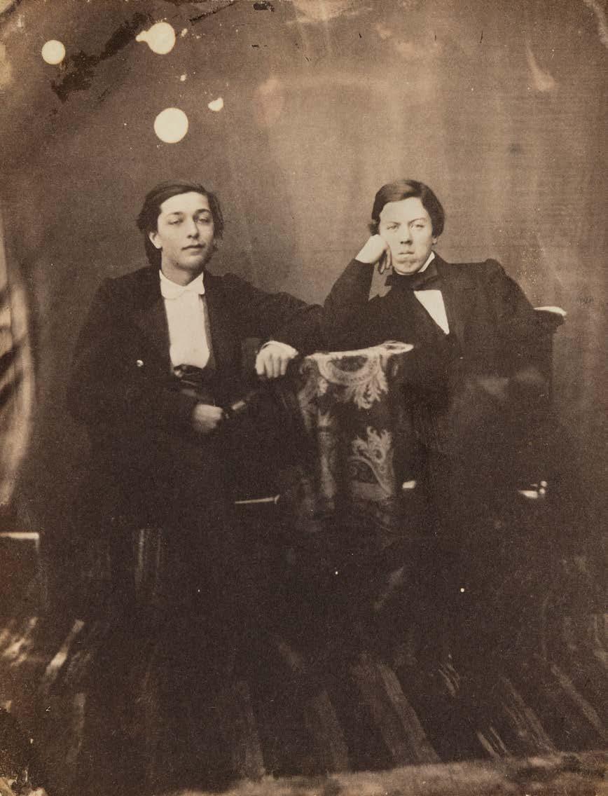 Kaksi nuorta miestä istumassa yhteismuotokuvassa. Pikkupöytää peittävä liina ja jalkojen alla oleva räsymatto toistuvat monessa Reinbergin otoksessa.