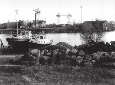 Mansikkasaaren telakka-alue Palosaaren suunnasta. Valokuva Gunnar Bäckman. Teoksesta Brändö - en stadsdel med själ. 1970 matala puurakenteinen sosiaalitila- ja portinvartinvartijanrakennus.