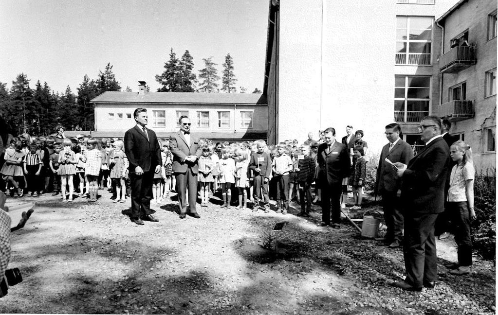 Ylhäällä: Kotikuusen istutustilaisuus Vuoksenniskan kansakoulun pihalla 30.5.1967 (kuva: Imatran kaupungin keskusarkisto). Vasemmalla: kuusia samalla kohtaa vuonna 2016.