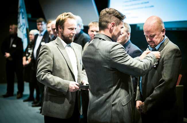 Puheenjohtaja Keskinen kiinnittää yhdistyksen ansiomerkkiä Mikko Ojasen takkiin asiamiehen seuratessa toimenpidettä.