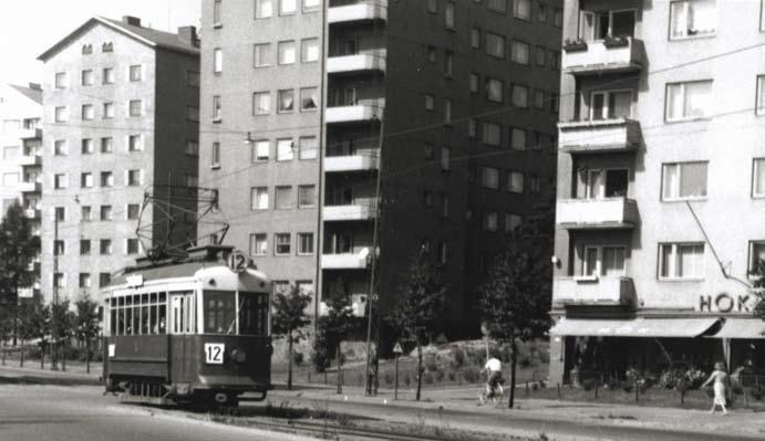 Perävaunun takana olevassa vuonna 1938 valmistuneessa kivitalossa saunottaa lähiseudun asukkaita vielä Aallokko Saunat. Fordson pakettiauto rek.nro BA 821 on juuri saapunut risteykseen Elimäenkadulta.