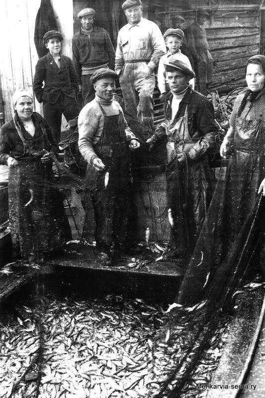 7 Silakkaverkkojen pudistelu on parhaillaan käynnissä Leo Salmen rannassa 1930-luvun loppupuolella. Tapahtumaa on seuraamassa muita kalastajia ja heidän lapsiaan.