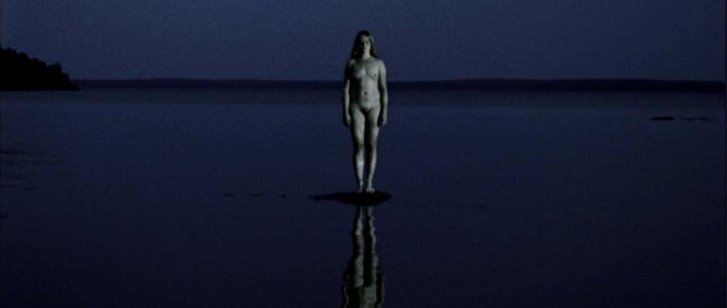 Kuva 13. Anni seisoo alasti järvellä kohtauksessa 24. 24 EXT. JÄRVI. ILTA. ANNI. Anni seisoo karilla keskellä järveä. Hän on alasti ja hänen ihonsa on kalpea. Kaikki on täysin tyyntä ja hiljaista.