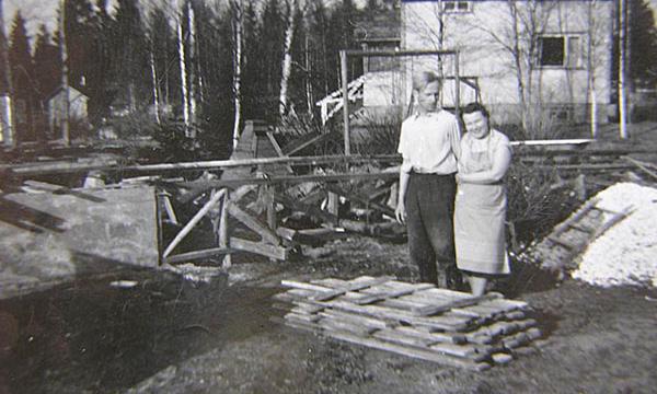Isä ja Äiti katsastavat tyytyväisinä isän puisia mökinsiirtokiskoja. Kohta saapuu vinssi, jolla entinen kesämökki siirretään saunaksi uuden kivijalan päälle.